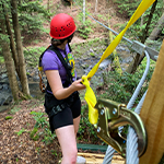 a girl standing on a zip-line platform overlooking a stream and pine trees